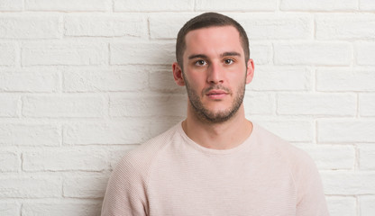 Young caucasian man standing over white brick wall with a confident expression on smart face thinking serious