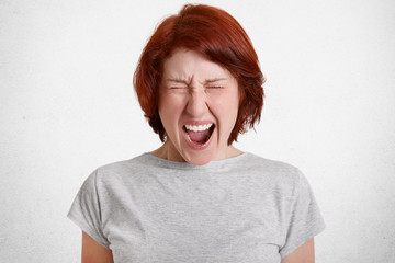 Devastated aggressive female with foxy hair screams desperately, has discontent facial expression, expresses aggression, exclaims loudly, dressed in casual grey t shirt, stands against white wall