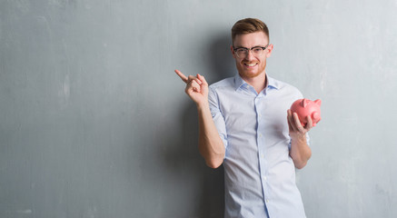 Young redhead man over grey grunge wall holding piggy bank very happy pointing with hand and finger to the side