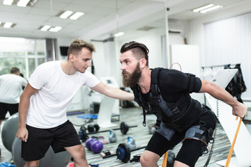 Young bearded man doing exercises in electrical muscular stimulation suit with her personal trainer at rehabilitation center.