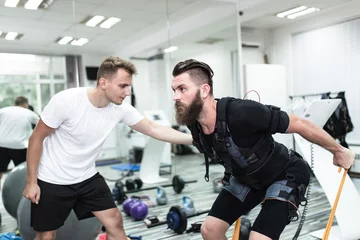 Tuinposter Young bearded man doing exercises in electrical muscular stimulation suit with her personal trainer at rehabilitation center. © hedgehog94