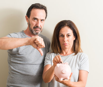 Middle age couple, woman and man, holding piggy bank with angry face, negative sign showing dislike with thumbs down, rejection concept