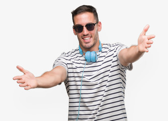 Naklejka premium Handsome young man wearing headphones looking at the camera smiling with open arms for hug. Cheerful expression embracing happiness.