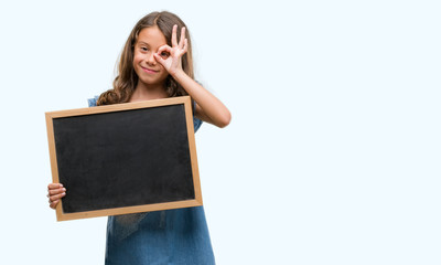 Brunette hispanic girl holding blackboard with happy face smiling doing ok sign with hand on eye looking through fingers