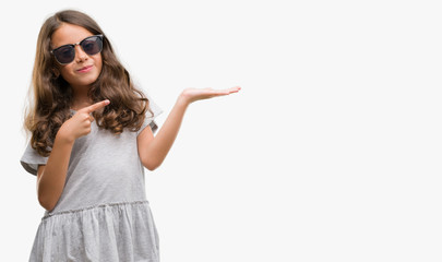 Brunette hispanic girl wearing sunglasses amazed and smiling to the camera while presenting with hand and pointing with finger.