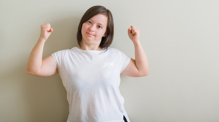 Down syndrome woman standing over wall showing arms muscles smiling proud. Fitness concept.