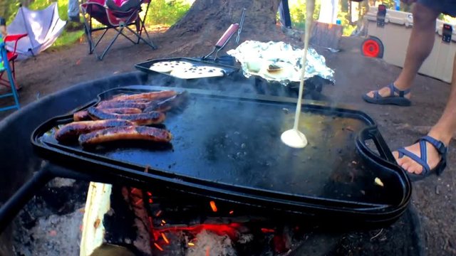 a slow motion, close up shot of making breakfast while camping. Sausage cooking while pouring pancake batter on the skillet and dropping in blueberries