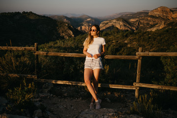 Cute girl in the T-shirt and shorts with sunglasses enjoying near the wood fence with a lot of green plants with green canyon on the background on the sunset in Spain. Traveler in the mountains.