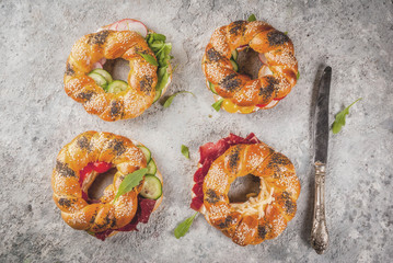 Variety of homemade bagels sandwiches with sesame and poppy seeds, cream cheese,  ham, radish, arugula, cherry tomatoes, cucumbers, white grey textured background copy space above