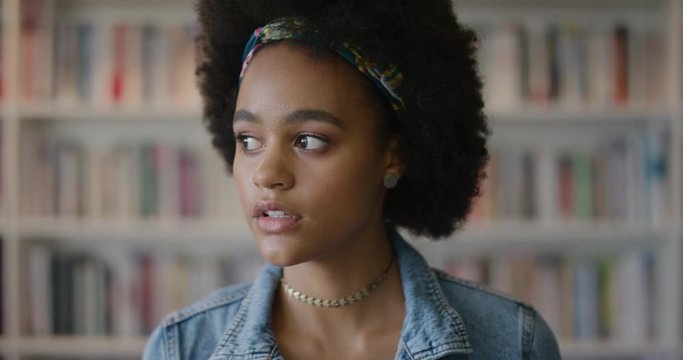 portrait beautiful young african american woman student looking around pensive contemplative smiling happy with afro hairstyle slow motion bookshelf background