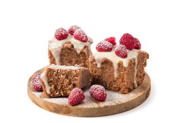 The chocolate cake decorated by white glaze and fresh berries of raspberry is strewed with icing sugar lies on a wooden round support. Separately on a white background. Horizontal shot. The place for 