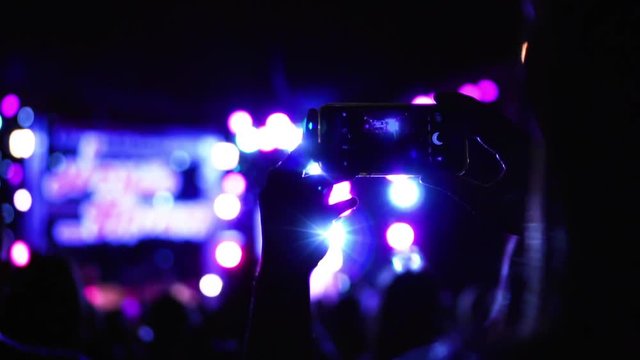 Asian woman person recording video with her phone in concert party with crazy flashing lights show and band on stage.