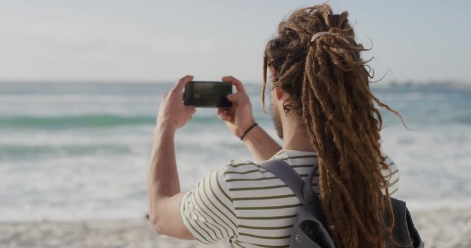 young tourist man taking photo on beach using smartphone camera technology of beautiful scenic ocean background caucasian male enjoying summer vacation travel 