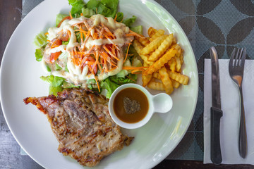 Top view on medium grilled homemade pork steak cooked by black pepper and salt serve with gravy sauce, fresh salad and french fries on white dish.