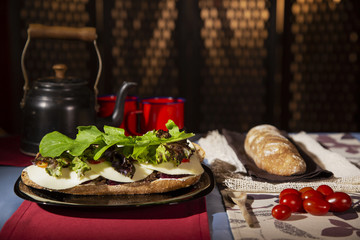 Open ciabatta bread sandwich with meat in a decorated table with cherry tomatoes. Teapot and cups in the background.