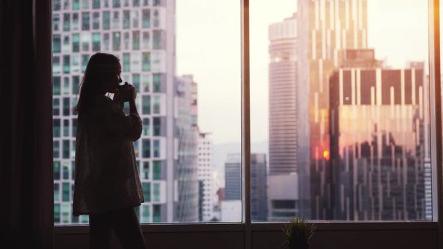 Beautiful womanstands by the window drinking coffee looks amazing cityscape view during beautiful sunset. slow motion. 3840x2160