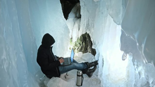Hacker hacks the server. Man to program on laptop in ice cave. Around the mysterious ice grotto. Man hides himself in a hood. Hacker has criminal cases on the Internet. Guy is warming himself with the