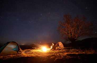 Night camping in mountains. Tourist man sitting between two illuminated tents on grassy valley, enjoying brightly burning campfire. Beautiful dark blue starry sky and big tree on the background