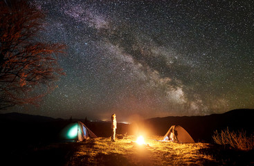 Peaceful camping night in mountains. Young tourist long-haired girl stands between two tents at burning campfire watching deep dark sky with lot of bright sparkling stars. Tourism and travel concept.