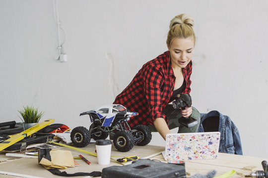 Attractive young delighted woman in red checkered shirt standing at wooden workbench with radio-controlled car holding remote controller and working on laptop placed nearby