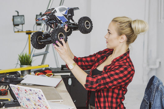 Side view of gorgeous blond female in casual clothes standing at wooden workbench holding toy radio-controlled car and looking attentively at it