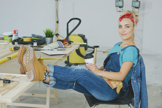 Young woman in overalls and red headband sitting comfortably on chair with legs on top of working desk full of instruments and tools holding paper cup with beverage and smiling looking at camera.