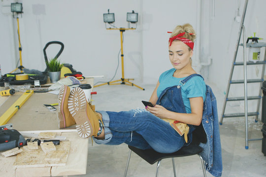 Beautiful young female in jeans overalls and red headband sitting relaxed with legs on desk with tools and using mobile phone in hand