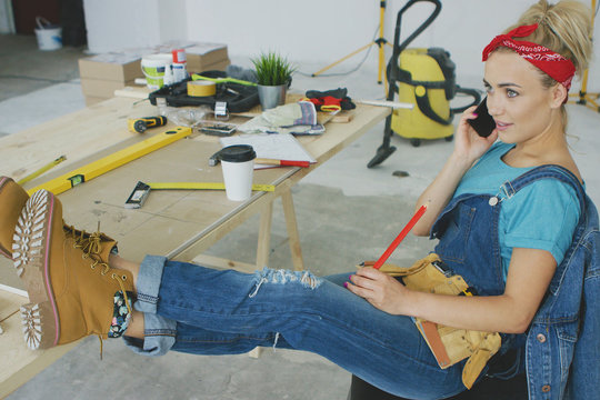 Relaxed young blond woman in jeans overalls sitting relaxed with legs on workbench with instruments talking on mobile phone and looking away