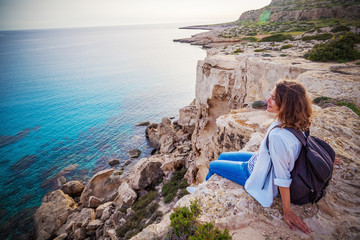 A stylish young woman traveler watches a beautiful sunset on the rocks on the beach, Cyprus, Cape...
