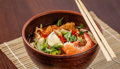 Asian Oriental Cuisine. Japanese spicy soup with seafood, shrimps, vegetables, lime, parsley, and hot pepper, in a plate of red clay, stands on a table in a restaurant. Copy space, selective focus