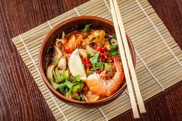 Asian Oriental Cuisine. Japanese spicy soup with seafood, shrimps, vegetables, lime, parsley, and hot pepper, in a plate of red clay, stands on a table in a restaurant. Copy space, selective focus