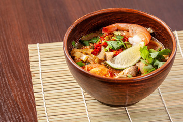 Asian Oriental Cuisine. Japanese spicy soup with seafood, shrimps, vegetables, lime, parsley, and hot pepper, in a plate of red clay, stands on a table in a restaurant. Copy space, selective focus