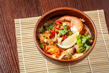 Asian Oriental Cuisine. Japanese spicy soup with seafood, shrimps, vegetables, lime, parsley, and hot pepper, in a plate of red clay, stands on a table in a restaurant. Copy space, selective focus