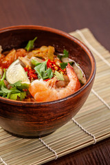Asian Oriental Cuisine. Japanese spicy soup with seafood, shrimps, vegetables, lime, parsley, and hot pepper, in a plate of red clay, stands on a table in a restaurant. Copy space, selective focus