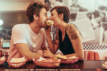Coupe in romantic mood sharing a burger at a restaurant - obrazy, fototapety, plakaty