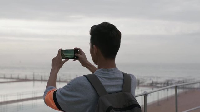 portrait of young asian man taking photo of cloudy seaside using phone