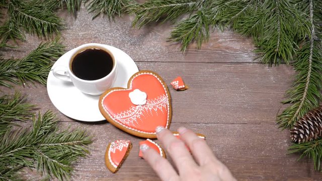 Food art.Broken heart. Fir tree, a cup of fresh-brewed coffee and heart-shaped gingersnaps placed on wooden background.one cookie is broken into pieces Top view.Female hands takes a piece.