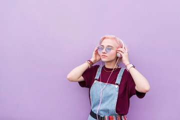 Stylish girl with pink hair and sunglasses brings music to the headphones on a purple background. Portrait of teenage girl listening to music in pink headphones.