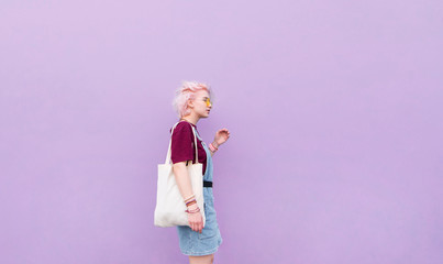 Stylish girl walking around the blue wall with a shopping bag. Girl with pink hair, glasses and shopper on the background of a purple wall