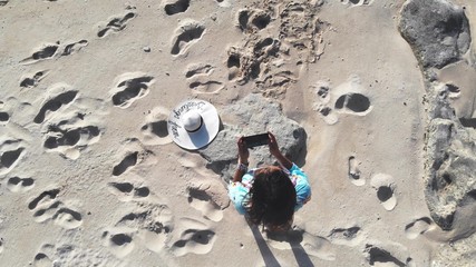 Top of young brunette pretty woman controls piloting drone copter using console holding phone at ocean beach. Smart girl operates fly quadcopter.