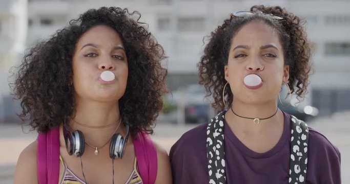 portrait attractive hispanic twin sisters blowing bubblegum enjoying summer vacation together smiling cheerful siblings in sunny urban city slow motion