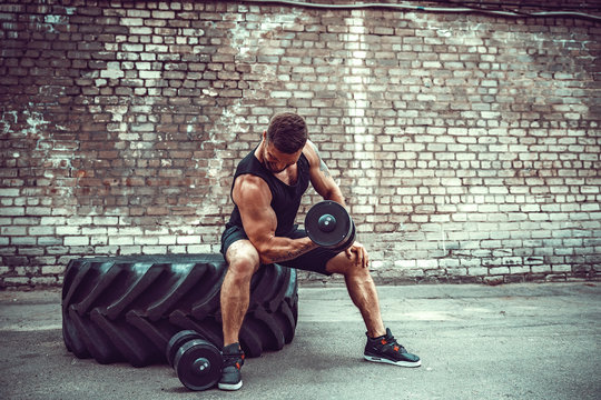 Athletic Man Working Out With A Dumbbell In Front Of Brick Wall While Sit On Big Tire. Strength And Motivation. Outdoor Workout. Exercise For The Muscles Biceps