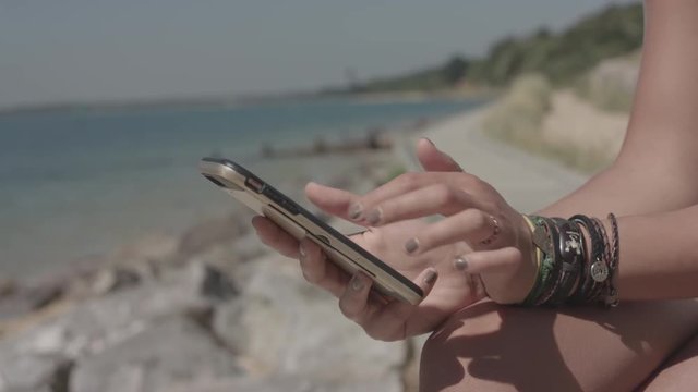 Young Attractive Woman Sat On The Beach Uses Her Smart Phone To Make Online Purchase - Ungraded