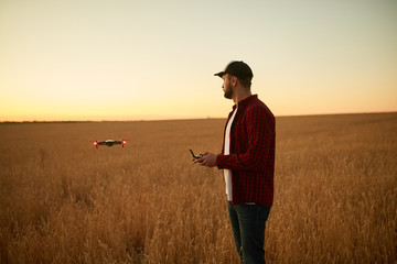 Farmer holds remote controller with his hands while quadcopter is flying on background. Drone hovers behind the agronomist in wheat field. Agricultural new technologies and innovations. Back view