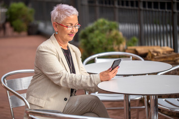 a fashion elderly woman is talking by skype wit a friend at the street coffee shop. side view...