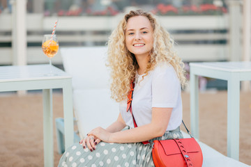 People and recreation concept. Lovely young female with curly bushy hair, wears t shirt and skirt, has bright small bag, rests outdoor, drinks fresh cocktail, looks positively directly at camera