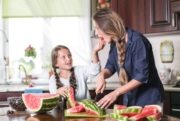 Cute little girl and her beautiful mom are cutting fruits, red watermelon and smiling while cooking in kitchen at home. Happy Family