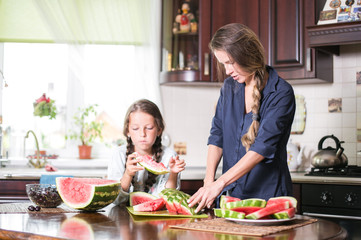 Cute little girl and her beautiful mom are cutting fruits, red watermelon and smiling while cooking in kitchen at home. Happy Family