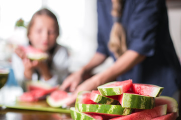 Cute little girl and her beautiful mom are cutting fruits, red watermelon and smiling while cooking in kitchen at home. Happy Family