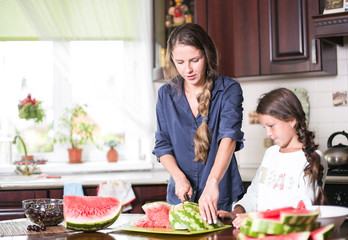Cute little girl and her beautiful mom are cutting fruits, red watermelon and smiling while cooking in kitchen at home. Happy Family